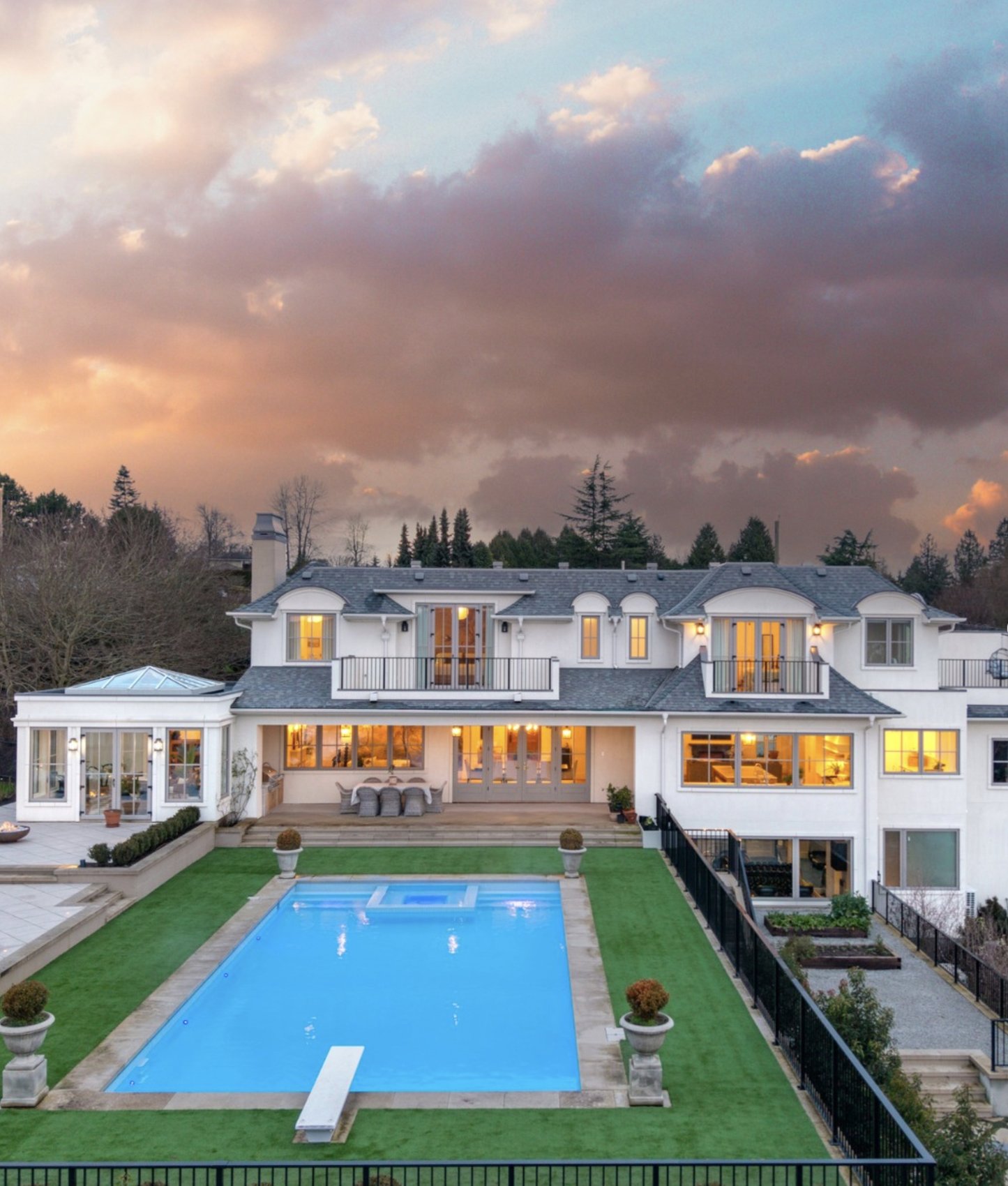 A grand white estate with manicured lawns and a pool beneath a dramatic sunset sky
